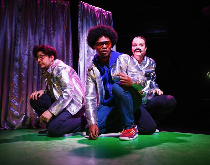 Photo of three cast members performing a scene, crouched in a group and if investigating something and trying to remain hidden.