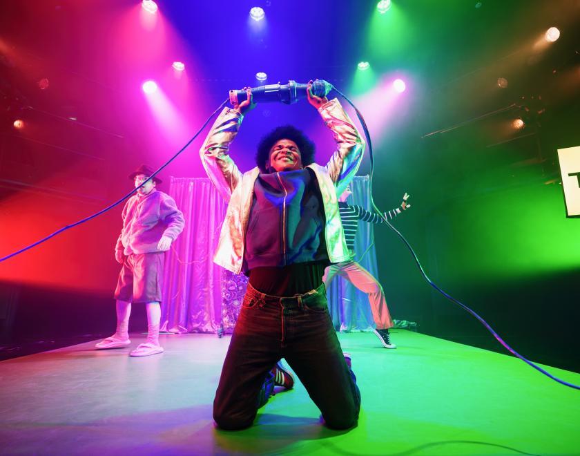 Photo of a performance of 'The Shivers'. Three cast members on a stage lit with bright colours.