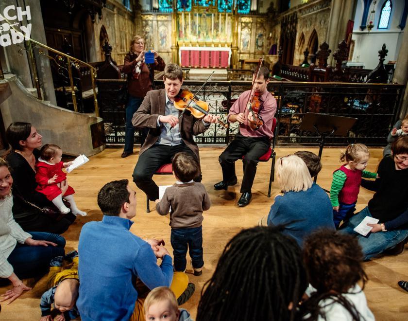 families and young children gather around two violinists