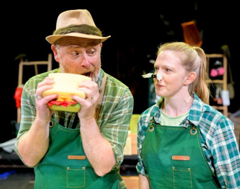 Photo of two actors performing 'Four Seasons. They are wearing green dungarees and green checked shirts, one is holding a huge prop sandwich and is about to take a bite, the other has an oversized prop bee or wasp on their nose. Some parts of the set are out of focus but visible behind them, and look to be made of wooden frames with plantpots and props hanging off them.