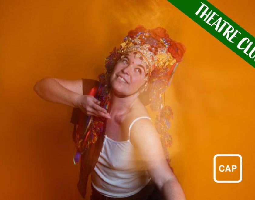 Photograph of performer Katherina Radeva, wearing a white vest top and elaborate gold and red headdress decorated with jewelled flowers. She is mid-dance, holding one hand up to her face and looking upwards. She is standing in front of an orange backdrop, and there is a blurred camera effect across the image, giving the impression of movement and reflection. White text reading 'Theatre Club' is overlaid across the top right corner and the symbol for Captioned Performance is in the bottom right corner