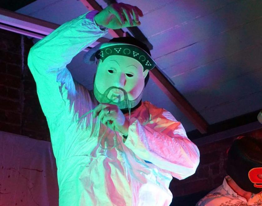 Person in a mask and white top staring at the camera, back lit in purples and pinks