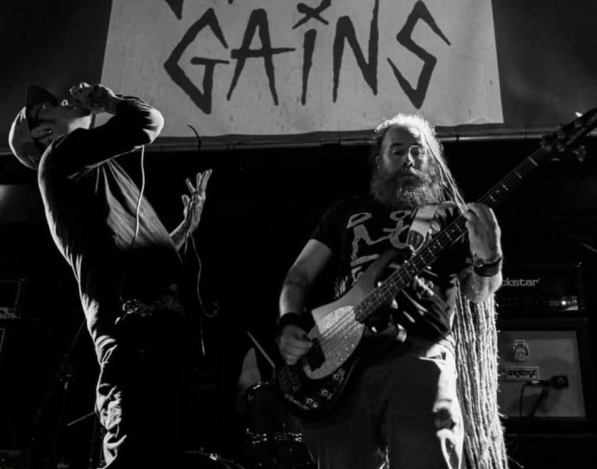 Black and white photo of two musicians one is singing the other is playing guitar. "Gotten Gains" can be read on a sign behind them.