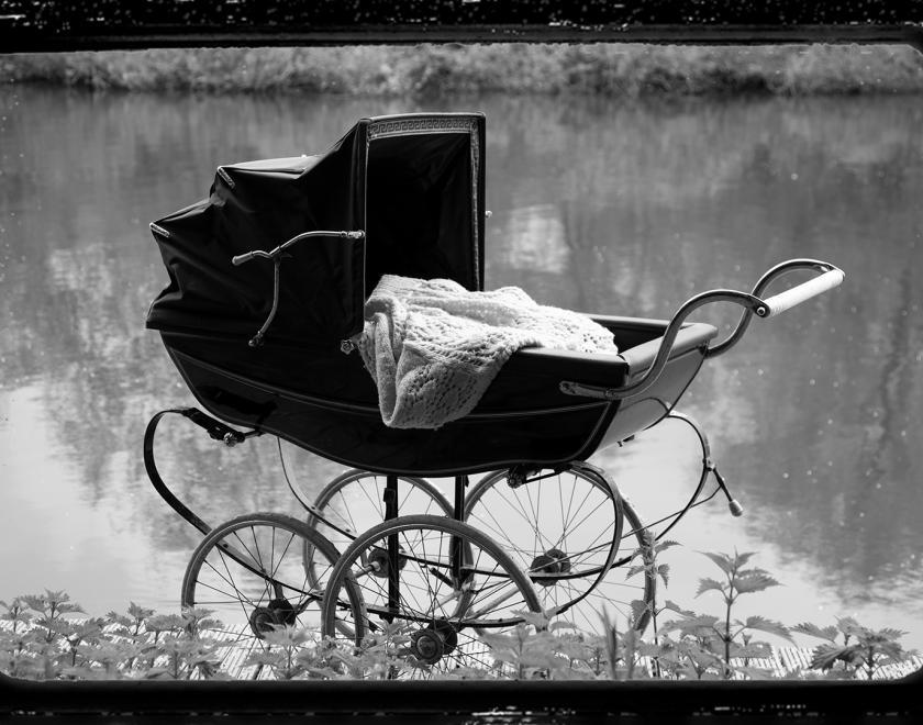 Black and white photo of an old fashioned babies' pram, empty, next to a river. A celluloid film frame has bee overlaid onto the image to make it look cinematic. 
