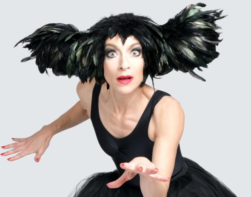 Performer Sarah-Louise Young, a white woman in her 40s, dressed as Kate Bush. She is wearing a black leotard and black tutu, and an elaborate headpiece made of black feathers. She is leaning forwards, holding both hands out in front of her with fingers outstretched, and wears a wide-eyed expression as if in surprise or fascination.