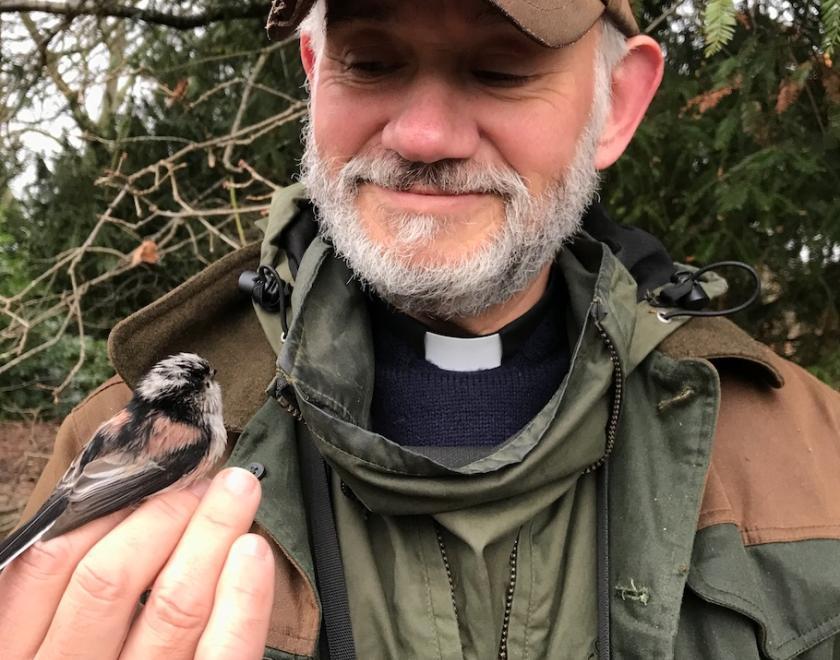 Andy Gosler in birding gear and clerical dog-collar holding a Long-tailed Tit