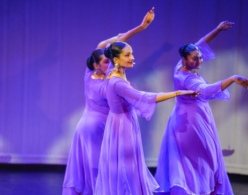 Three South Asian women in purple dresses performing classical dance poses