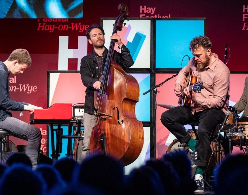 four musicians on stage. Left to right, they play electric piano, upright bass, electric guitar and drums.