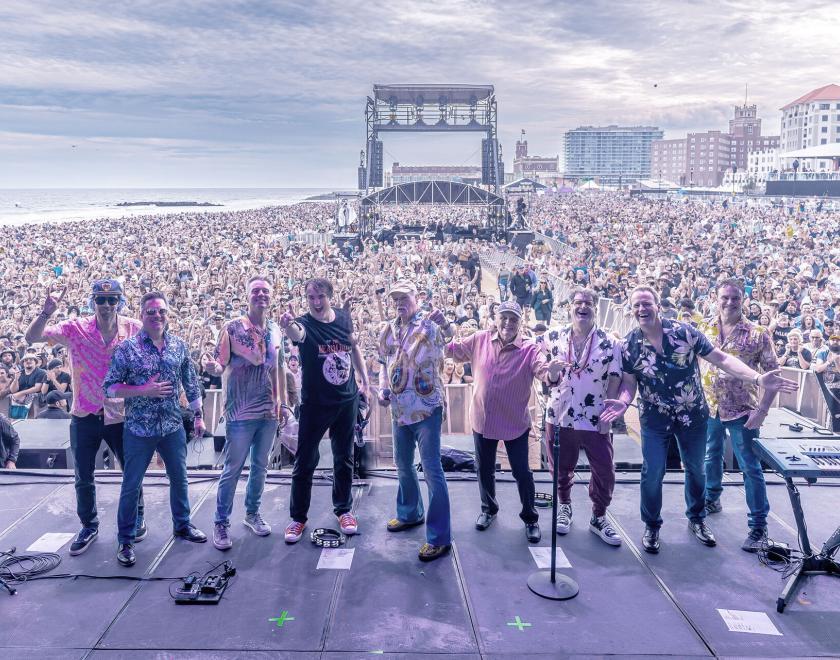 the beach boys band standing in a line on-stage. A massive crowd can be seen behind them.