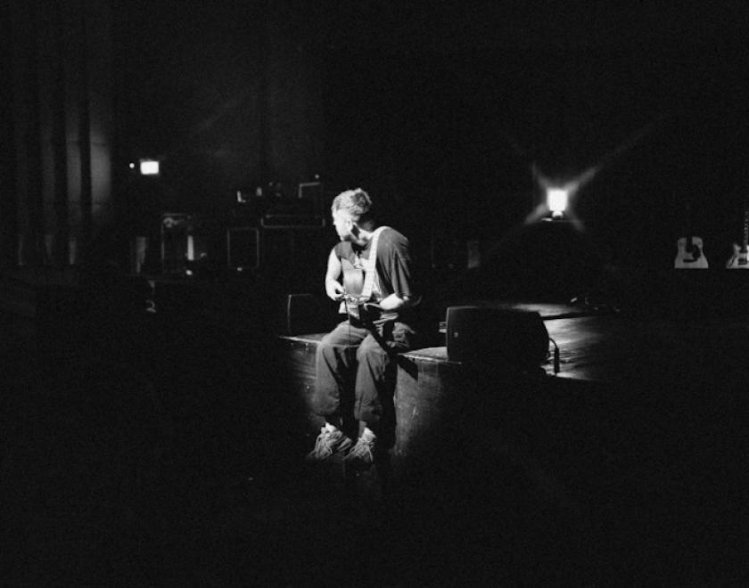 Black and white photo of Benjamin Francis Leftwich sitting on the edge of a stage with a guitar, facing away from the camera.