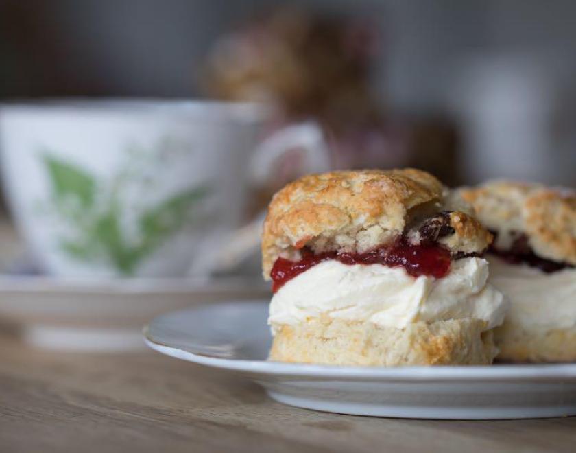 Scone with cream and jam on a plate, with a cup of tea in distance