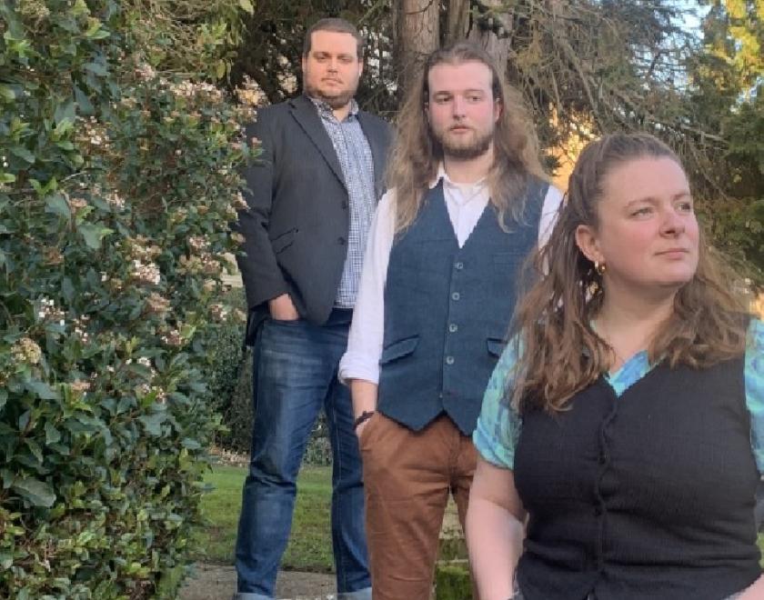 Image of the three members of Culverake, standing on stone steps, surrounded by trees and next to an ivy-clad wall. The trio are standing informally and are dressed in casual clothes.