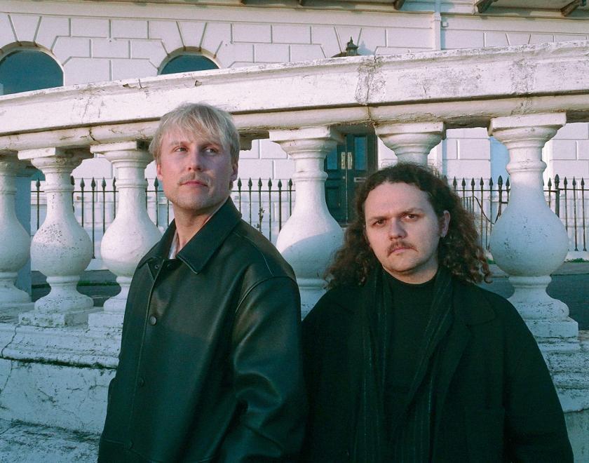 Puma Theory band members standing outside a stately home. One has short blonde hair and a blonde moustache, the other long brown hair and a dark moustache.