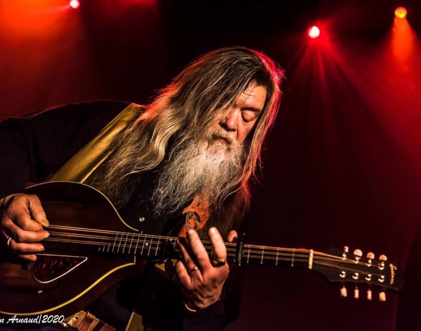 Person playing a mandolin. The background is dark but with patches illuminated by red stage lights. The musician is dressed in dark clothing and has long straight hair and a long white beard