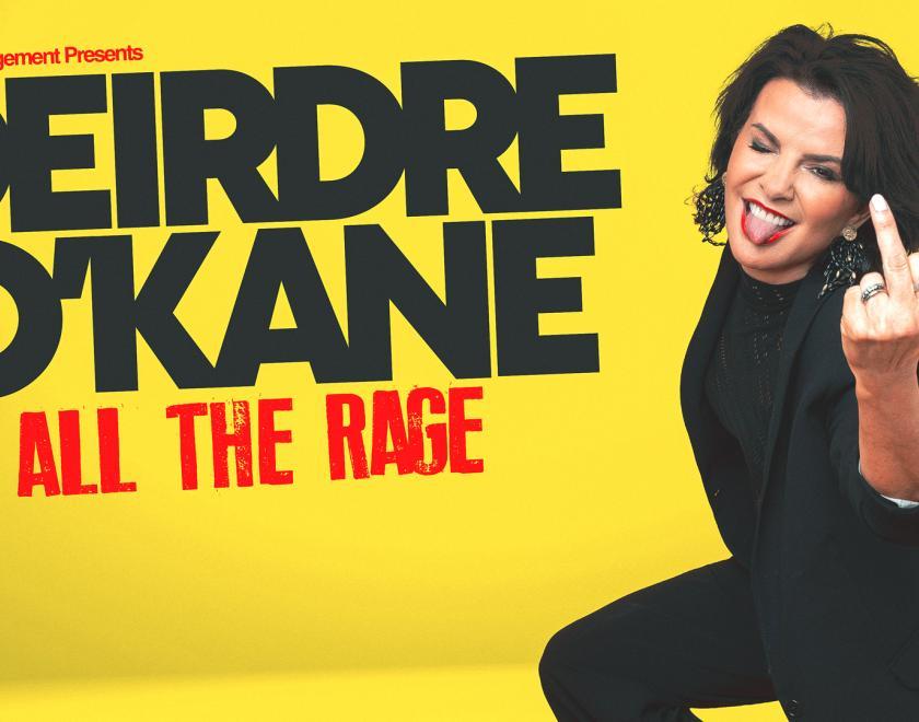 Deirdre O'Kane on a yellow background, smiling and holding up middle finger. 