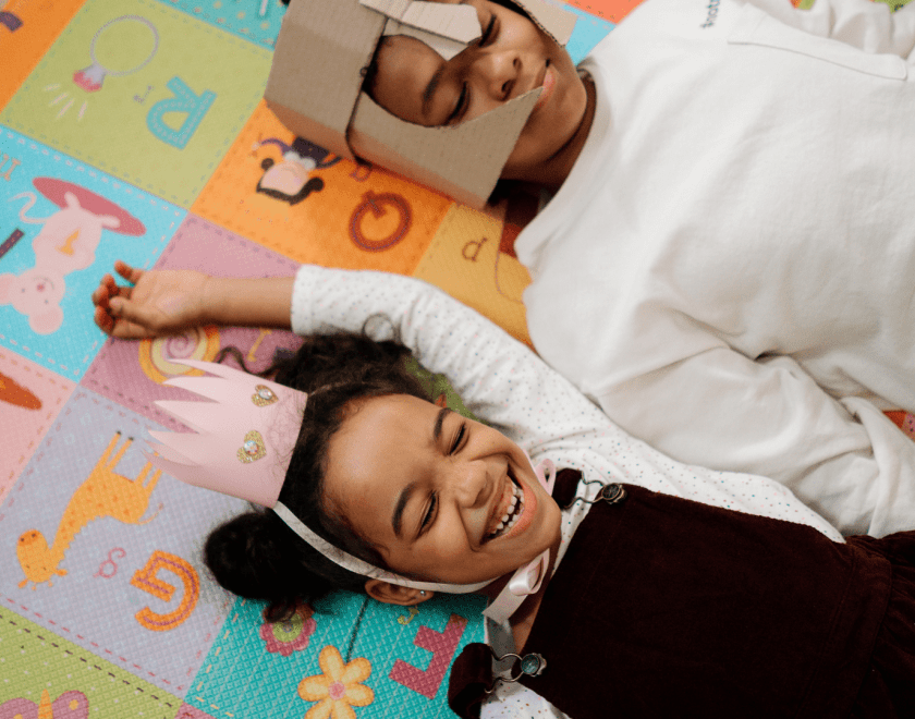 Two young children laying on the floor wearing cardboard crown and knights helmet