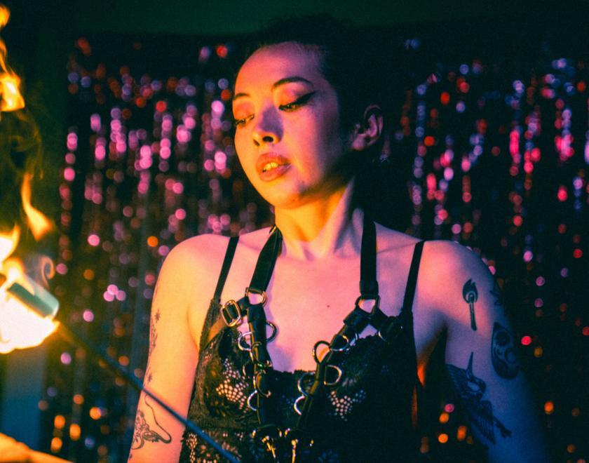 A tattooed, femme presenting performer poses in front of a shining purple glitter curtain. They wear a sultry black lace garment, and are holding flaming swords as part of a fire eating act
