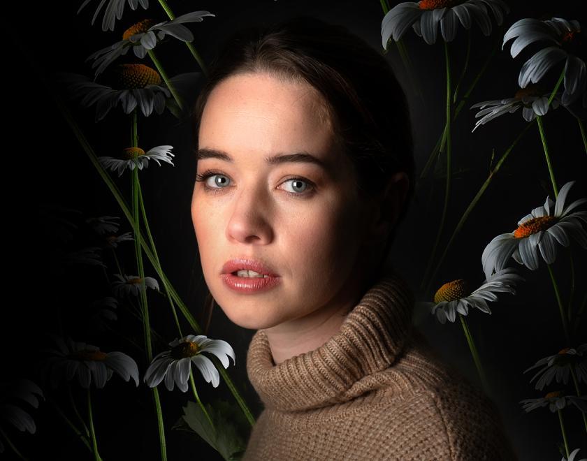 Anna Popplewell looking vacantly, behind her are flowers shrouded in a darkness