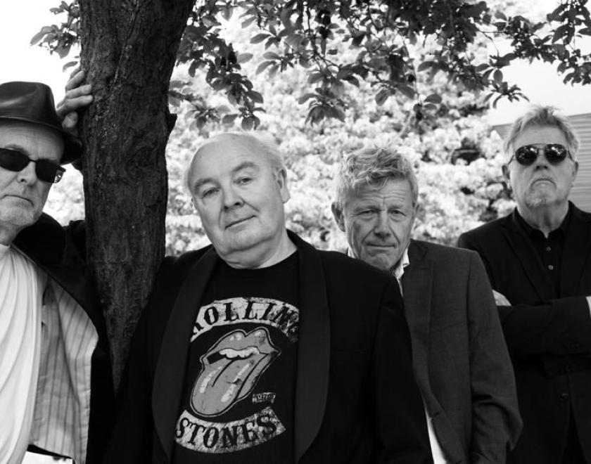 Black and white line up of 4 musicians all looking towards the camera and wearing dark jackets. The individual on the far left has dark glasses and a hat and leans on the trunk of a tree which shelters the whole band. The next person is bald and wears a Rolling Stones T-shirt, the third member of the band is slightly behind him and then the final member also wears dark glasses and has his arms crossed.