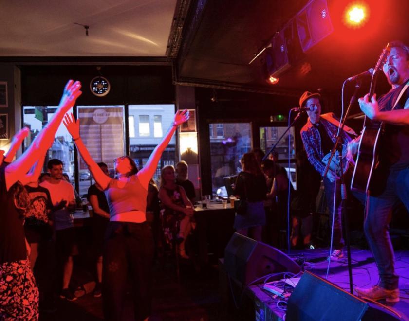 A pub gig; two musicians are on a stage on the right of the image, they are both wearing jeans, playing guitars and singing and bathed in violet light. Centre and left of the image shows the audience who are bathed in red light and have their arms in the air.