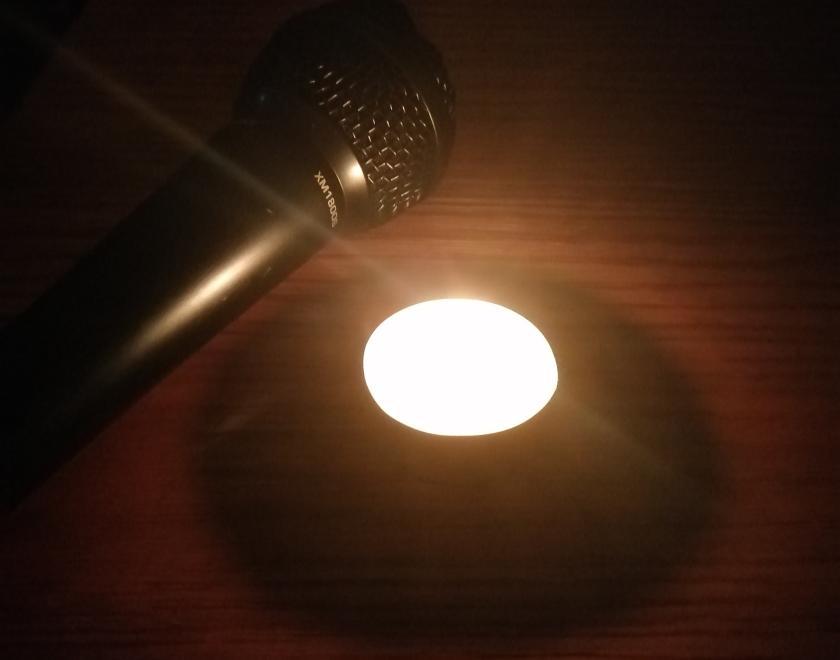 A tealight candle shines in a dark space illuminating a wooden surface; just on the edge of the pool of light is a black microphone