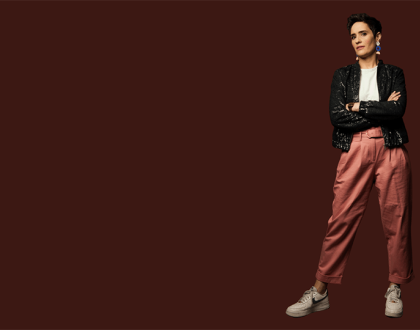 Photograph of Jen Brister, wearing a white t-shirt, black sparkly jacket, dusky pink trousers and white trainers. She is standing with her arms crossed against a dark red backdrop.