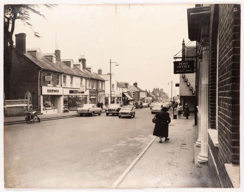 A high street with shops, cars and people walking