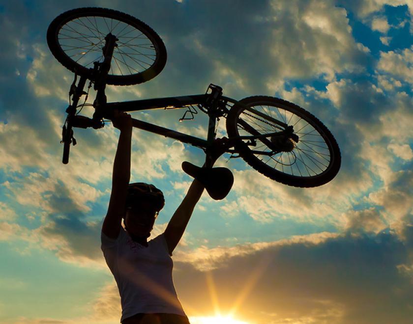 On Track promotional image. A silhouette of a woman holding a bicycle above her head with a sunset sky behind her