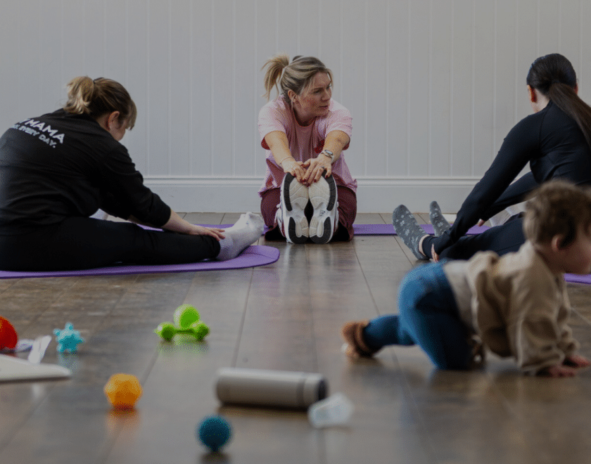 Parents taking part in Our Story & Co.’s Postnatal Move & Support group, enjoying gentle, postnatal-friendly stretching exercises on purple mats while a baby explores nearby toys. A relaxed, welcoming session combining movement, wellbeing and connection, where babies are welcome alongside their parents.