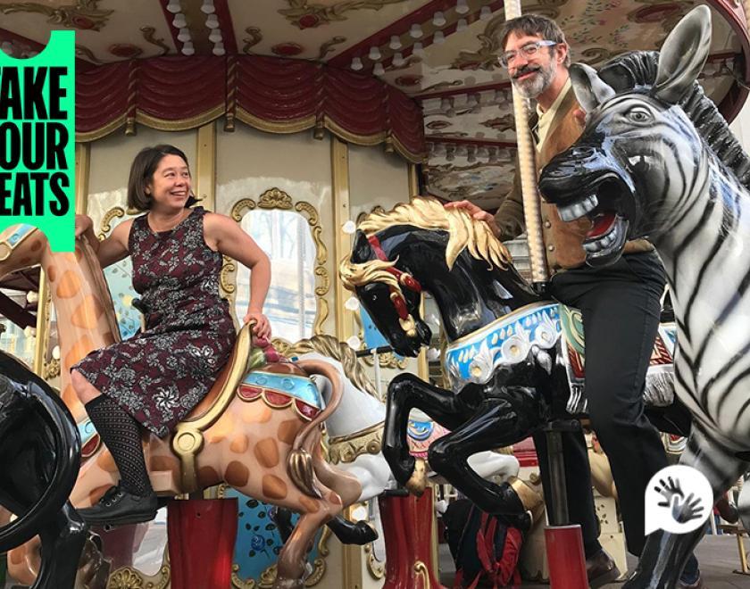 Photograph of performers Jo Fong and George Orange riding an old fashioned carousel, sitting on models of a giraffe and horse respectively. 'Take Your Seats' logo is in the top left corner and the British Sign Language symbol is in the bottom right corner.