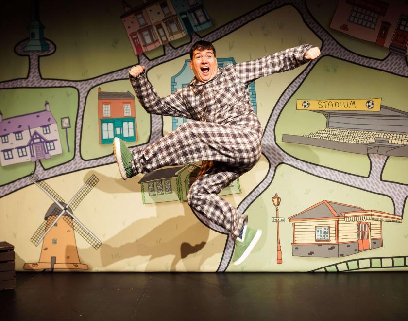 A smiling performer in checked pyjamas leaps energetically across the stage in front of a colourful illustrated map featuring houses, a football stadium, a windmill and village landmarks. The playful image captures the fun, adventurous spirit of a family theatre production.