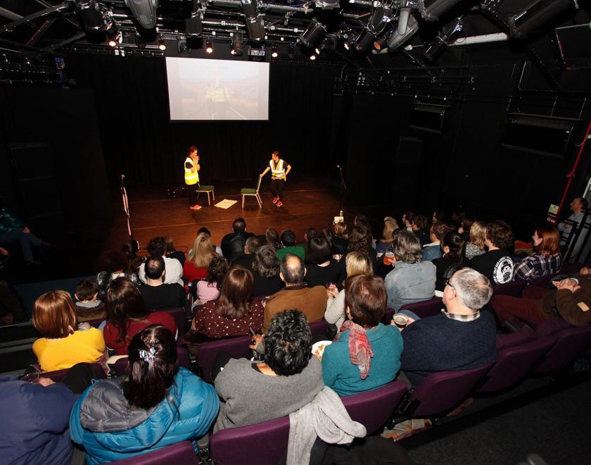 Photo taken from the back of the main space at South Street, with the backs of the audience visible and two performers in hi-vis jackets on stage.