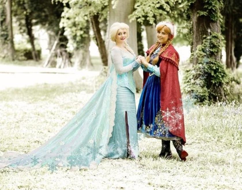 The image shows two women holding hands in a forest. One is dressed like Elsa and one is dressed like Ana from Frozen.