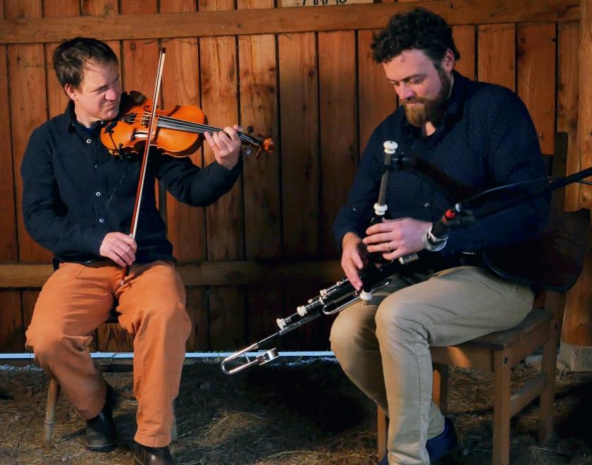 Photo of Bradon and Dominic, the two members of Straight from the Jug sitting on a bench with a wood-clad wall behind and playing fiddle and uileann pipes.