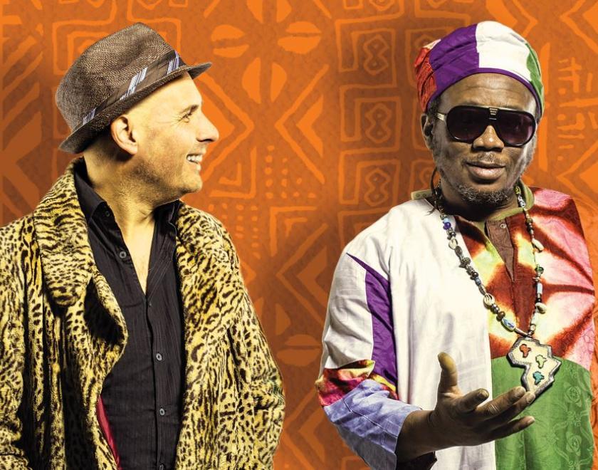 A patterned orange backdrop with Two musicians shown from the waist up.  The figure on the right looks towards us, he is black with a little bit of facial hair and is wearing large sunglasses and multi-coloured clothing. The figure on the left has turned his head to face his companion, he is white with stubble and wears a leopard print jacket.
