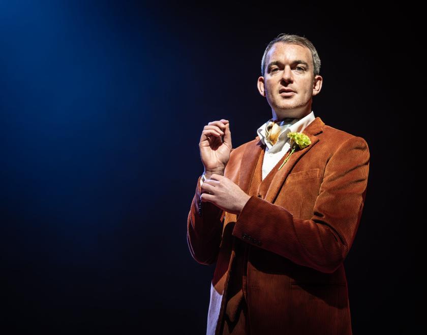 A man stands adjusting his sleeve cuff. wearing an orange suit against a dark blue background. 
