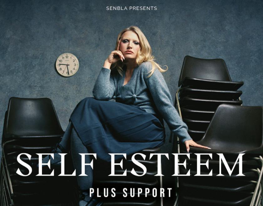 Singer Self Esteem wearing a blue blouse and skirt, sitting on a stacked pile of chairs
