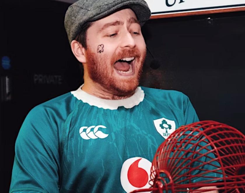 Man with a ginger beard and a big grin, wearing a flat cap and Ireland football shirt, holding a bingo wheel