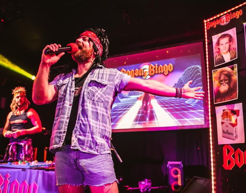 Two men in mullet wigs on stage with bingo game bits and pieces