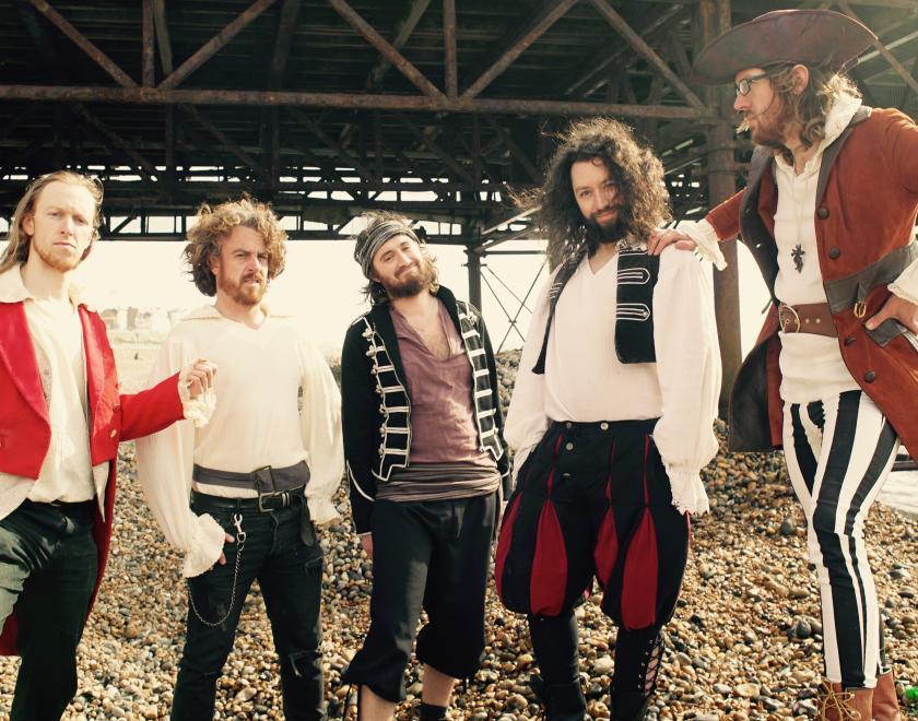 Five band members dressed in piratical garb stand on a pebbly beach with a large metal structure behind