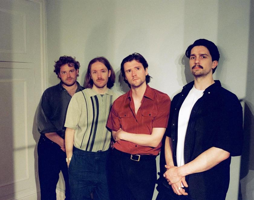 Members of the band "Do Nothing", four twenty-something men with hipster haircuts and moustaches, standing in front of a door