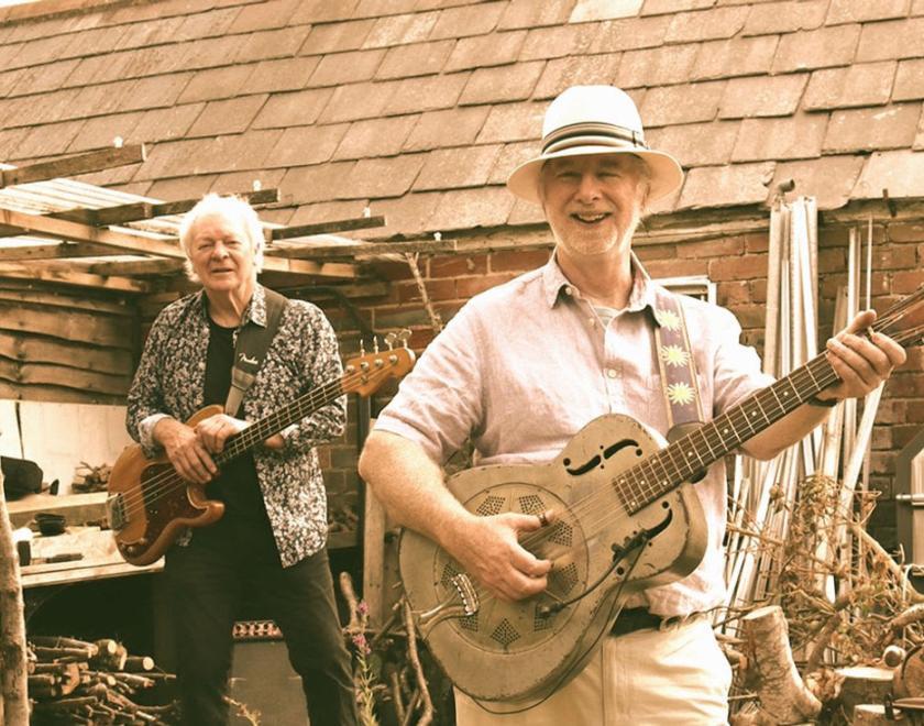 Four older white male musicians  outside a brick and slate building with firewood around. 1st and 3rd musicians in the row wear hats. Left to Right they play: Percussion, bass, National steel guitar, mandolin