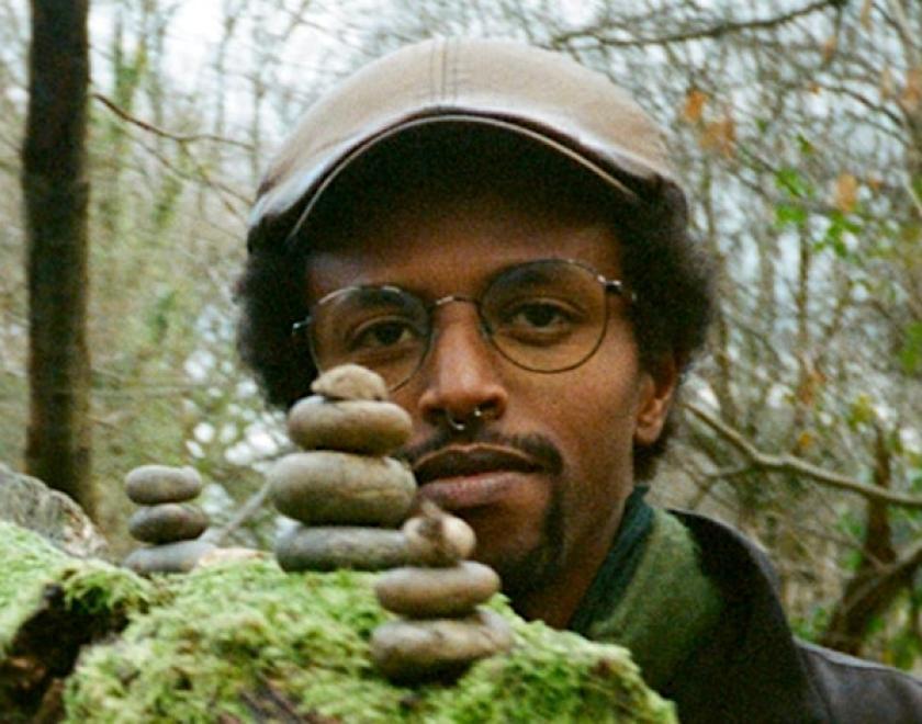 The head of  a dark skinned musician is visible behind a mossy log with piles of stones on it. The background is wooded