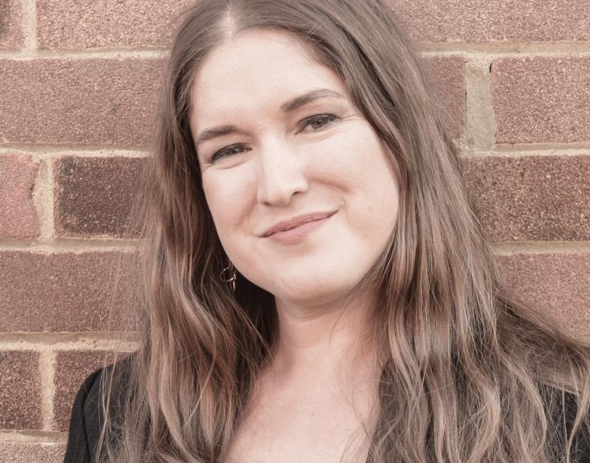 Singer Jessica Lee Morgan with long brown hair and a big smile, standing in front of a brick wall