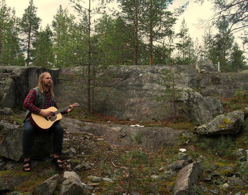 Long-haired musician wearing chequed flannel shirt, denim waistcoat, jeans and sandals with an acoustic guitar sits on a rock to the left of the image in front of a stony outcrop in pine wood.
