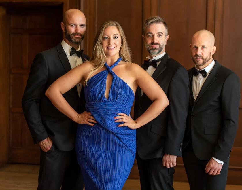 Three bearded men in tuxedos and a lady with blonde hair in a blue evening dress