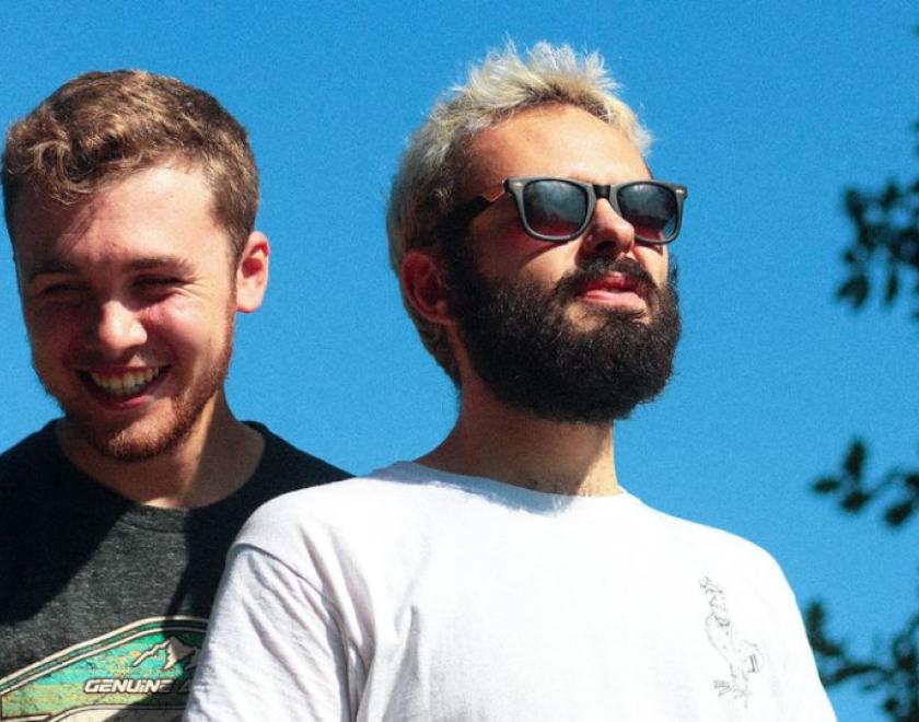 Two musicians are shown from the waist up with a strikingly blue sky behind them. The one on the left of the image wears a grey t-shirt, they have pale skin, short ash-brown hair, short thinnish facial hair and their eyes crinkle in a smile. The person on the right of the image is wearing a white t-shirt and sunglasses; they also have pale skin, their hair is short bleach blonde and they have a short dark beard
