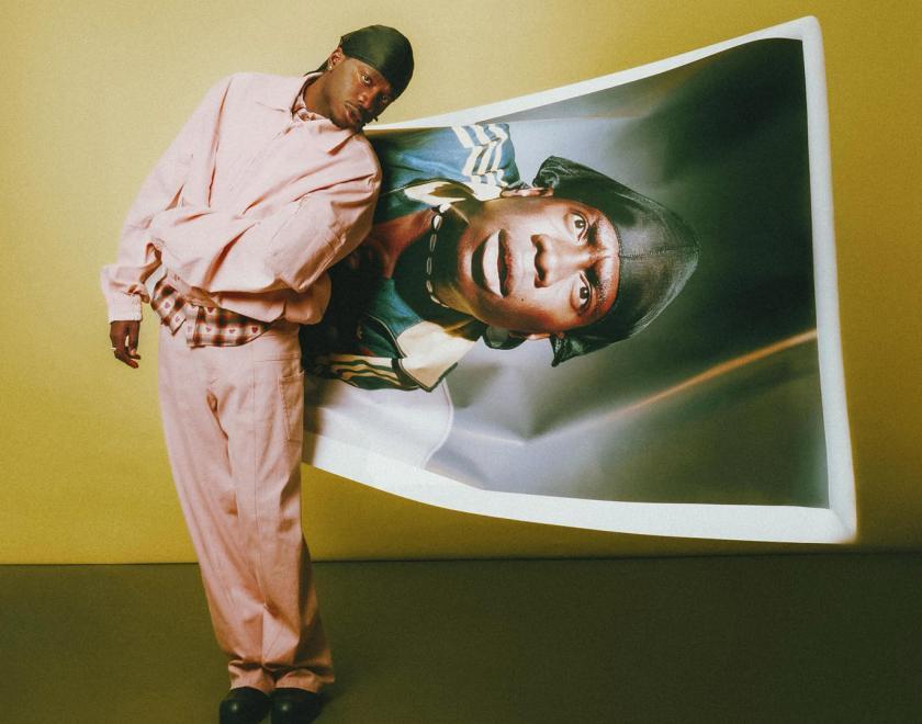 Image of singer Master Peace, a young black man in a pink suit, stnaidng next to a warped oversized photo of himself