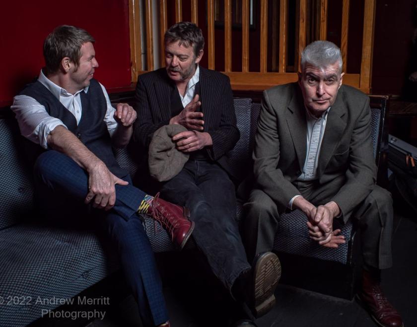 Three bandmates sit on a corner sofa. All are dressed smart casual with open neck shirts and a hint of rockabilly. The two on the left are chatting to each other and the one on the right is staring at the camera quizzically