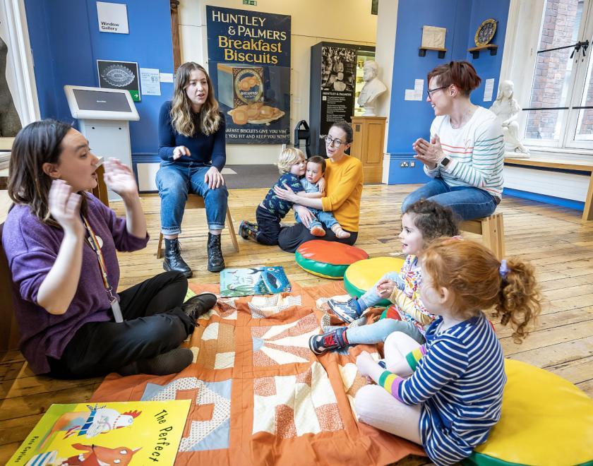 Rhymetime at Reading Museum, member if staff is sat on a patchwork quilt surrounded by toddlers and their parents